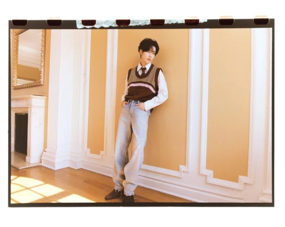 Portrait of an East Asian male in a sweater vest and jeans posing in a neutral-toned interior with ornate wall trim and sunlight.