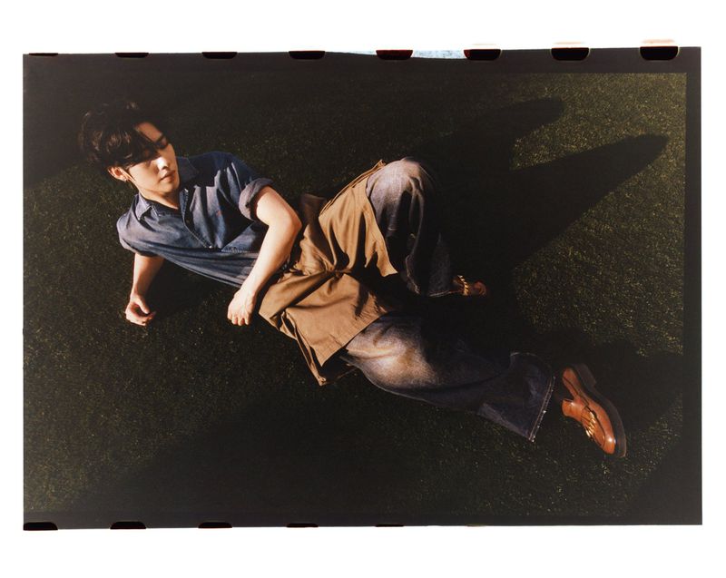 A young adult man lies on grass in blue shirt, brown shorts, jeans, and dress shoes, creating a casual yet stylish fashion portrait.
