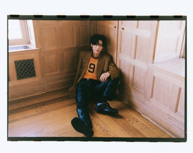 Young adult male with dark hair in a brown suit and orange shirt poses indoors, sitting on the wood floor in a corner.