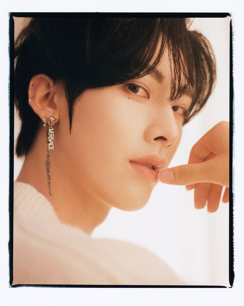 Portrait of a young man with dark hair, wearing a white sweater, gazing at the camera. He has a designer earring with the text 'VERSACE'.