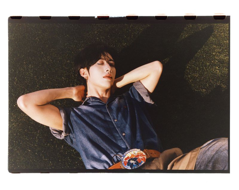 Person in a denim shirt lies in the grass with their hands behind their head and a large colorful belt buckle.