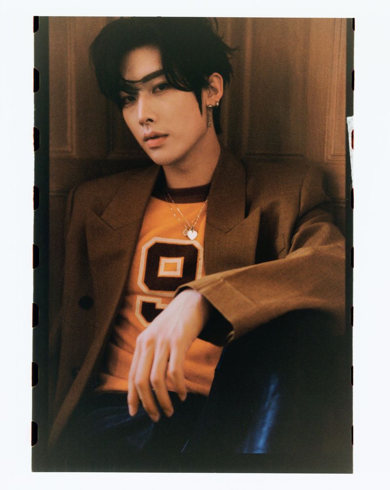 Portrait of a young adult with dark hair wearing a brown blazer and orange shirt with a large number 9, against a neutral backdrop.