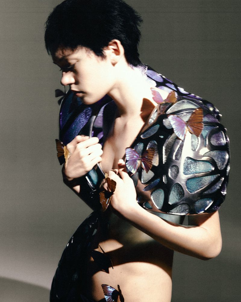 Fashionable young adult poses in a silver and blue butterfly-themed outfit against a plain background, shot in a studio.