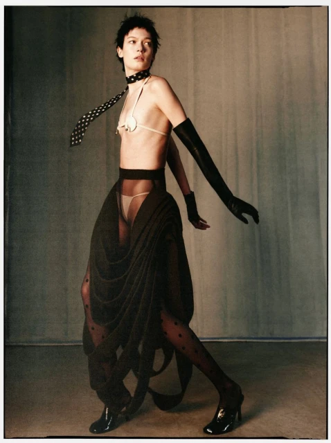 Woman in dramatic dark fashion, light skin, wearing black gloves, stockings, and heels, with a polka-dot tie and minimal bikini top, posing indoors.