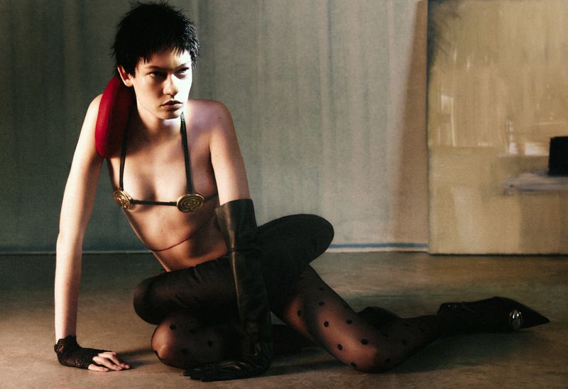 Woman in fashion pose with golden bra, tights, and gloves in an artistic setting with a neutral background.