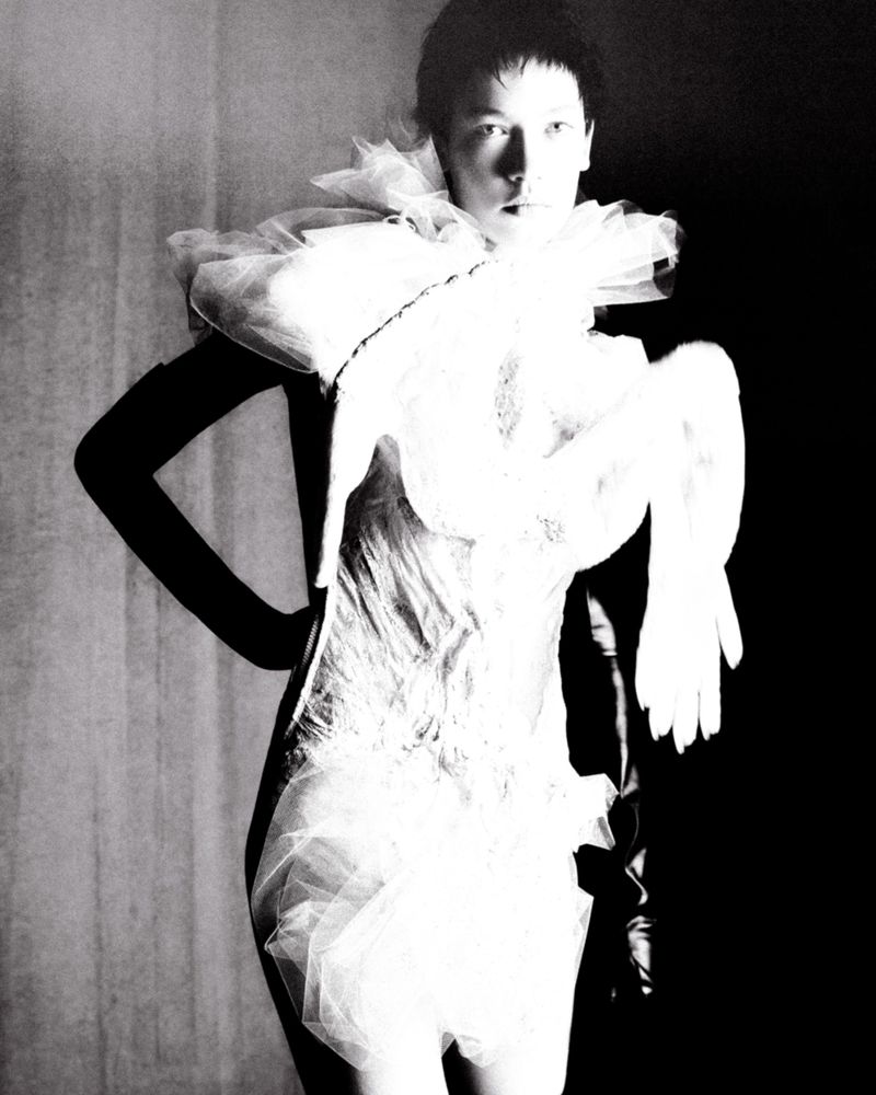 Monochrome fashion shot of a slender adult woman posing in a white tulle dress with theatrical sleeves against a dark background.