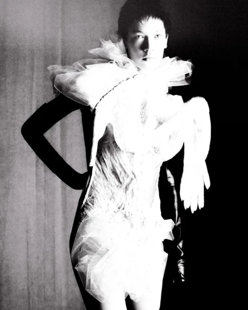 Monochrome fashion shot of a slender adult woman posing in a white tulle dress with theatrical sleeves against a dark background.