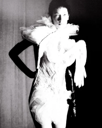 Monochrome fashion shot of a slender adult woman posing in a white tulle dress with theatrical sleeves against a dark background.