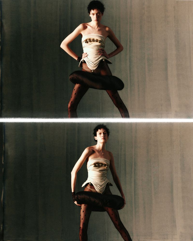 A woman poses in avant-garde fashion featuring a strapless corset, patterned tights, and a unique black tube around her hips.
