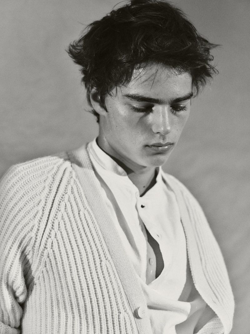 Monochrome portrait of a young adult male with tousled dark hair wearing a white button-down shirt and cardigan, gazing downward in soft light.