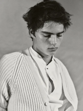 Monochrome portrait of a young adult male with tousled dark hair wearing a white button-down shirt and cardigan, gazing downward in soft light.