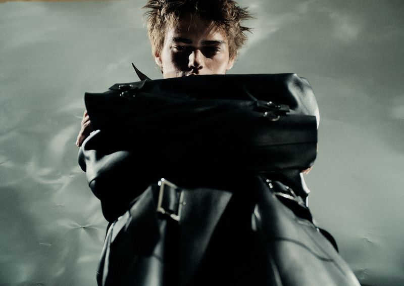 Fashion shot of an adult man wearing a leather jacket and standing indoors against a grey textured background, looking at the camera.
