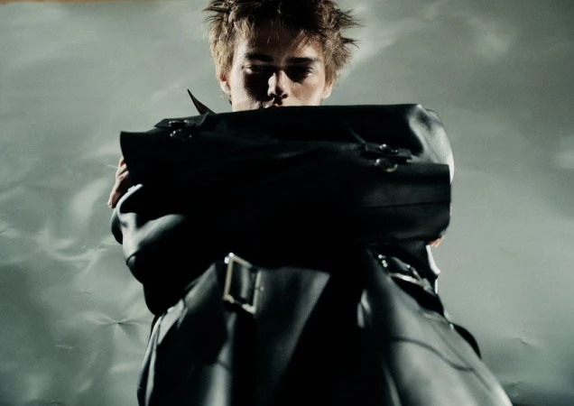 Fashion shot of an adult man wearing a leather jacket and standing indoors against a grey textured background, looking at the camera.