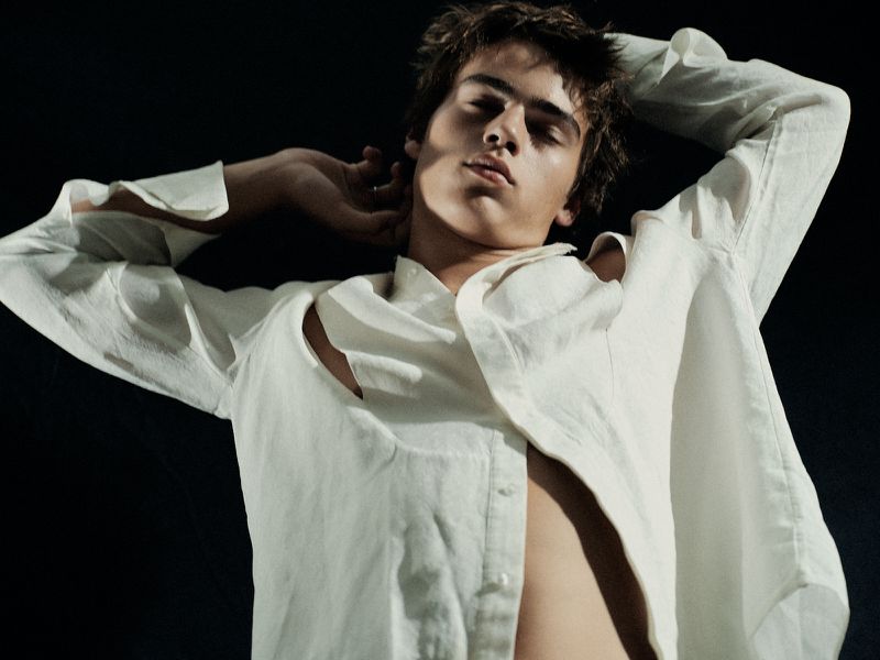 Portrait of a young adult male model with hands behind his head wearing a partially unbuttoned white linen shirt on a black background.