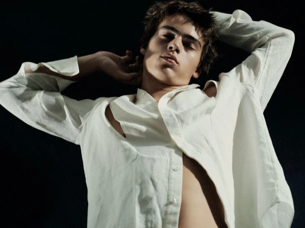 Portrait of a young adult male model with hands behind his head wearing a partially unbuttoned white linen shirt on a black background.