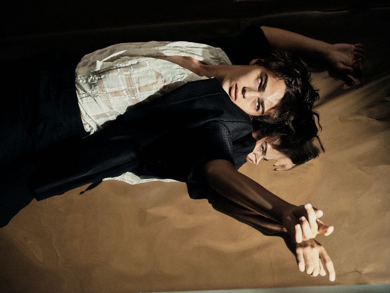 Two adults lie on a brown surface. The person in the foreground wears a dark top, the other person wears a light top. Dark lighting.