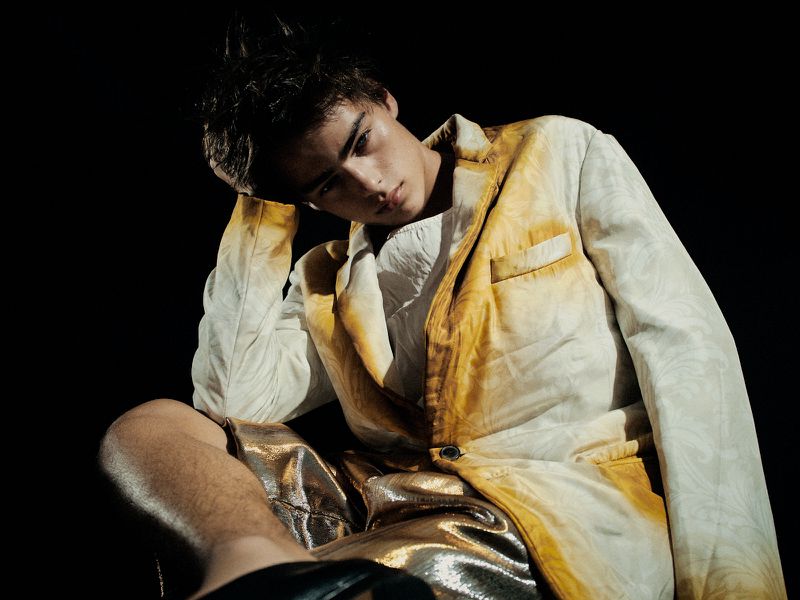 Man in a white patterned jacket with gold metallic shorts poses against a dark background, showcasing a unique fashion style.
