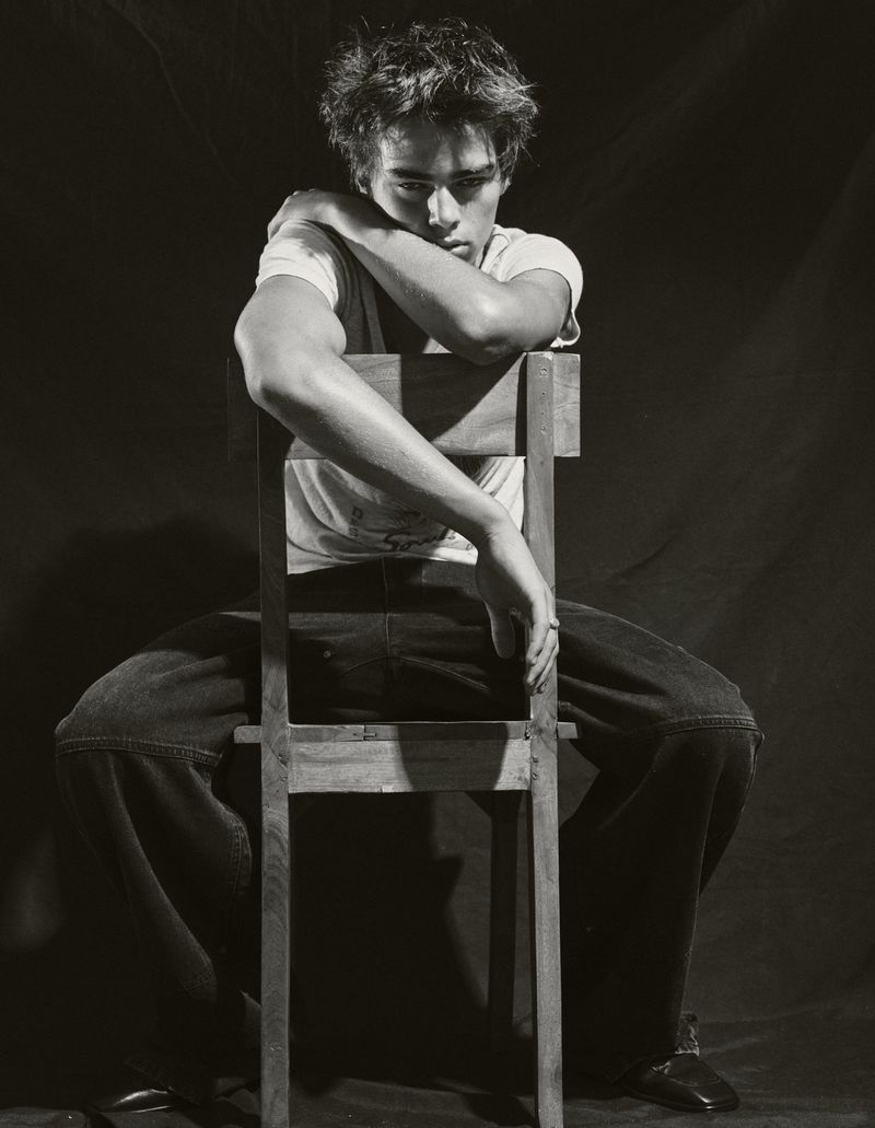 Monochromatic portrait of a young adult male sitting backwards on a wooden chair with arms crossed, wearing a white t-shirt and jeans.