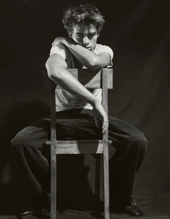 Monochromatic portrait of a young adult male sitting backwards on a wooden chair with arms crossed, wearing a white t-shirt and jeans.
