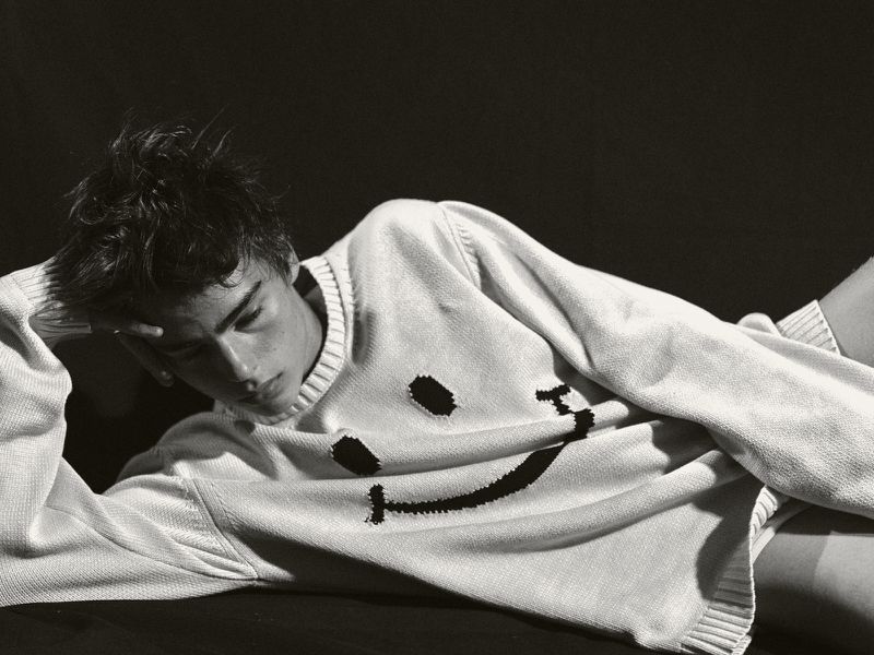 Monochrome portrait of a young man lying down in a white sweater with a smiley face design against a dark background.