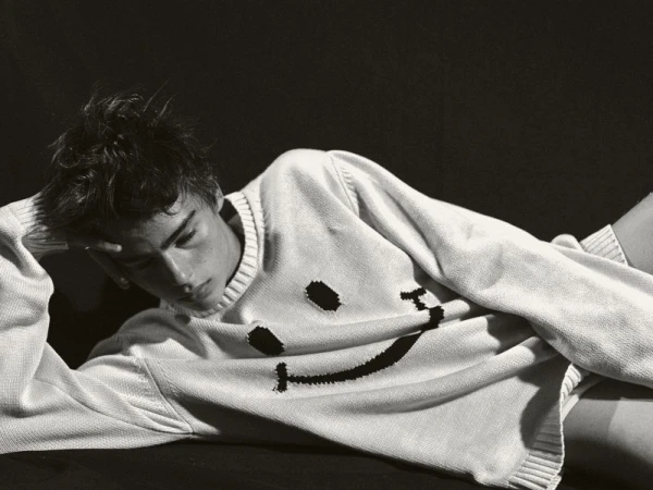 Monochrome portrait of a young man lying down in a white sweater with a smiley face design against a dark background.