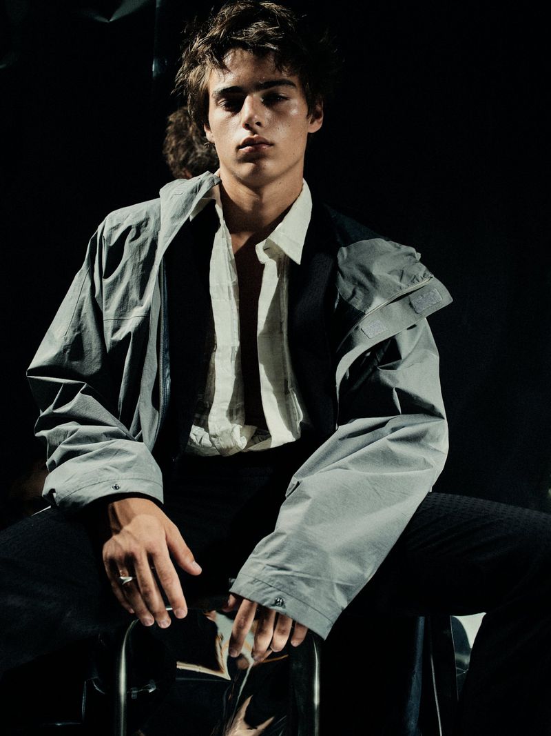 Fashion portrait of a young adult man in gray jacket and striped shirt sitting on a metal stool against a dark background.