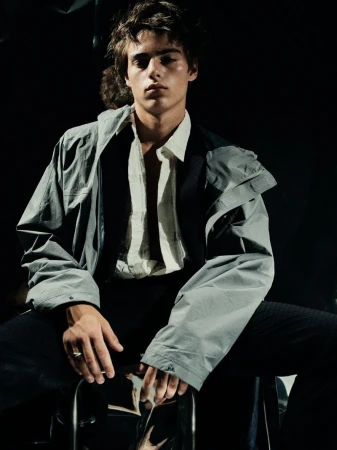 Fashion portrait of a young adult man in gray jacket and striped shirt sitting on a metal stool against a dark background.