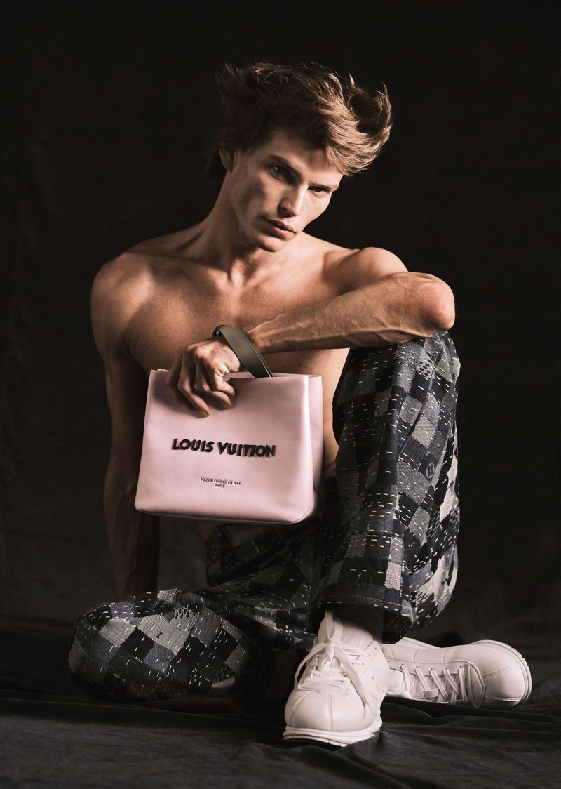 Androgynous male fashion model poses shirtless with a pink Louis Vuitton handbag against a dark background in a studio setting.