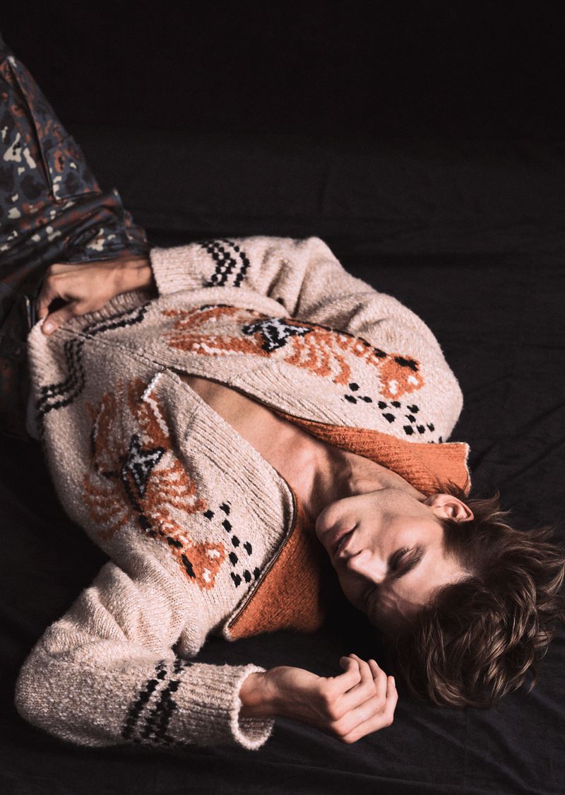 Adult male with light brown hair reclines on a black surface, wearing an open patterned knit sweater with printed dark pants.