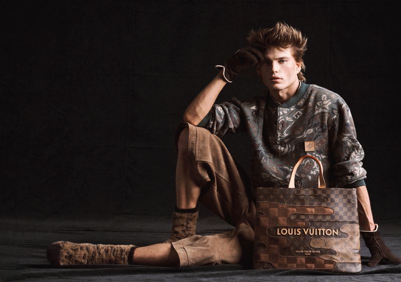 A male fashion model poses indoors next to a Louis Vuitton bag. He wears a patterned sweater, brown pants, and matching brown gloves.