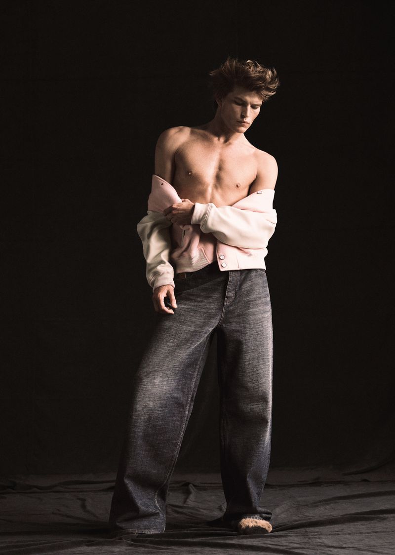 Fashionable young adult male poses shirtless in wide leg jeans and an off-the-shoulder jacket against a dark neutral backdrop.