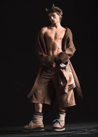 Full-shot of a male fashion model with an unbuttoned brown overcoat, matching brown shorts, and brown winter boots in a studio setting.