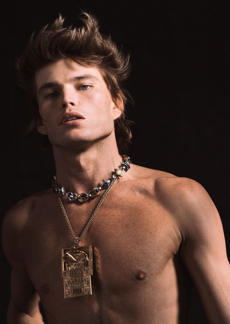 Male fashion model with bare chest and golden necklace posing in a photo studio with a dark background in a medium close-up shot.