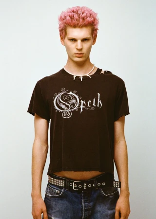 A young adult male poses in a studio wearing a black band t-shirt with pink hair and a studded belt. The photo has a clean editorial style.