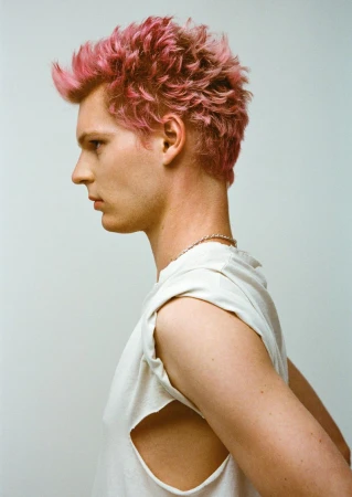 Profile portrait of a young adult male with vibrant pink hair, wearing a white sleeveless shirt against a clean, light grey backdrop.
