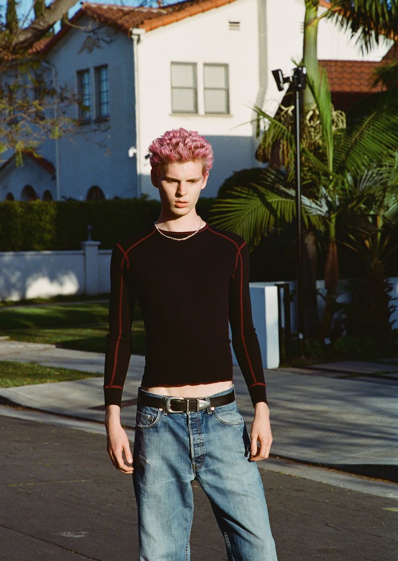 Young adult with pink hair stands on a paved road wearing a black long-sleeved shirt, jeans, and a belt with a silver buckle.