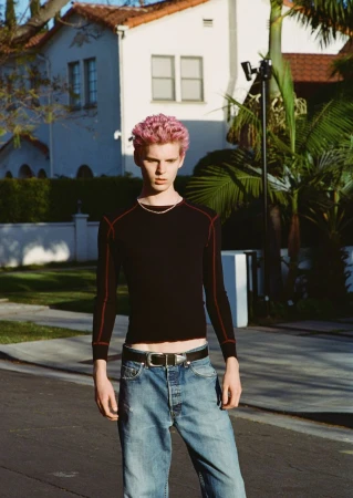 Young adult with pink hair stands on a paved road wearing a black long-sleeved shirt, jeans, and a belt with a silver buckle.