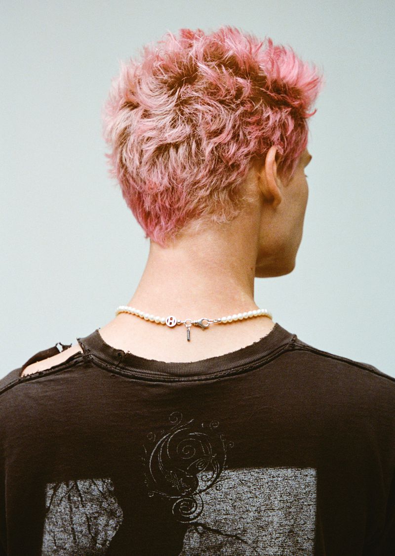 A person is seen from the rear, showing pink hair, wearing a pearl necklace and a black t-shirt with a graphic, against a blue background.