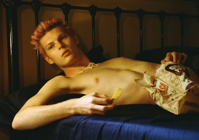 A young adult male with pink hair lies in bed, topless, eating chips from a bag in a dimly lit interior setting.