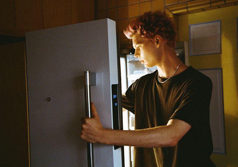 Young man with pink hair opens a modern grey refrigerator in an indoor kitchen setting. He is wearing a dark t-shirt and silver necklace.