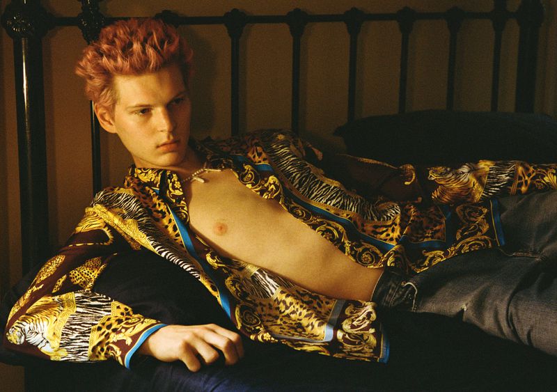 Adult male with pink hair poses in printed shirt, jeans, and necklace on a bed with dark headboard in an indoor setting.