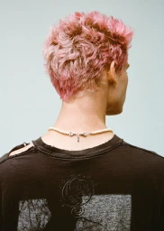 Close-up back view of a model with short pink hair wearing a pearl necklace and dark distressed t-shirt against a plain light background