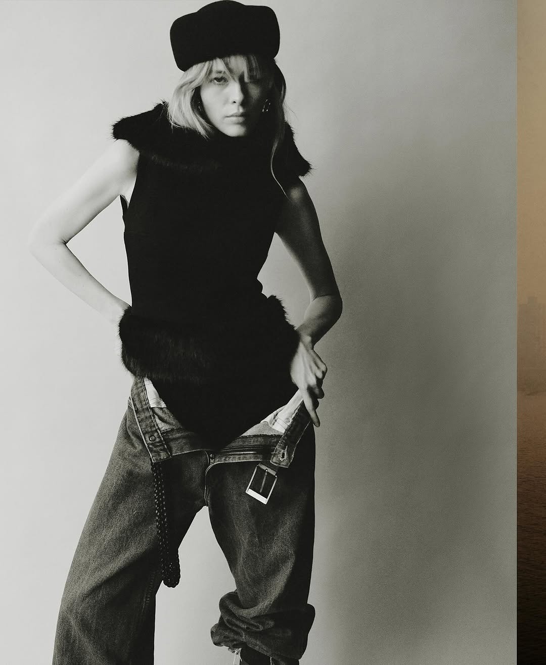 Adult female fashion model poses indoors in a black sleeveless top, baggy blue jeans, and a dark cap against a neutral backdrop.