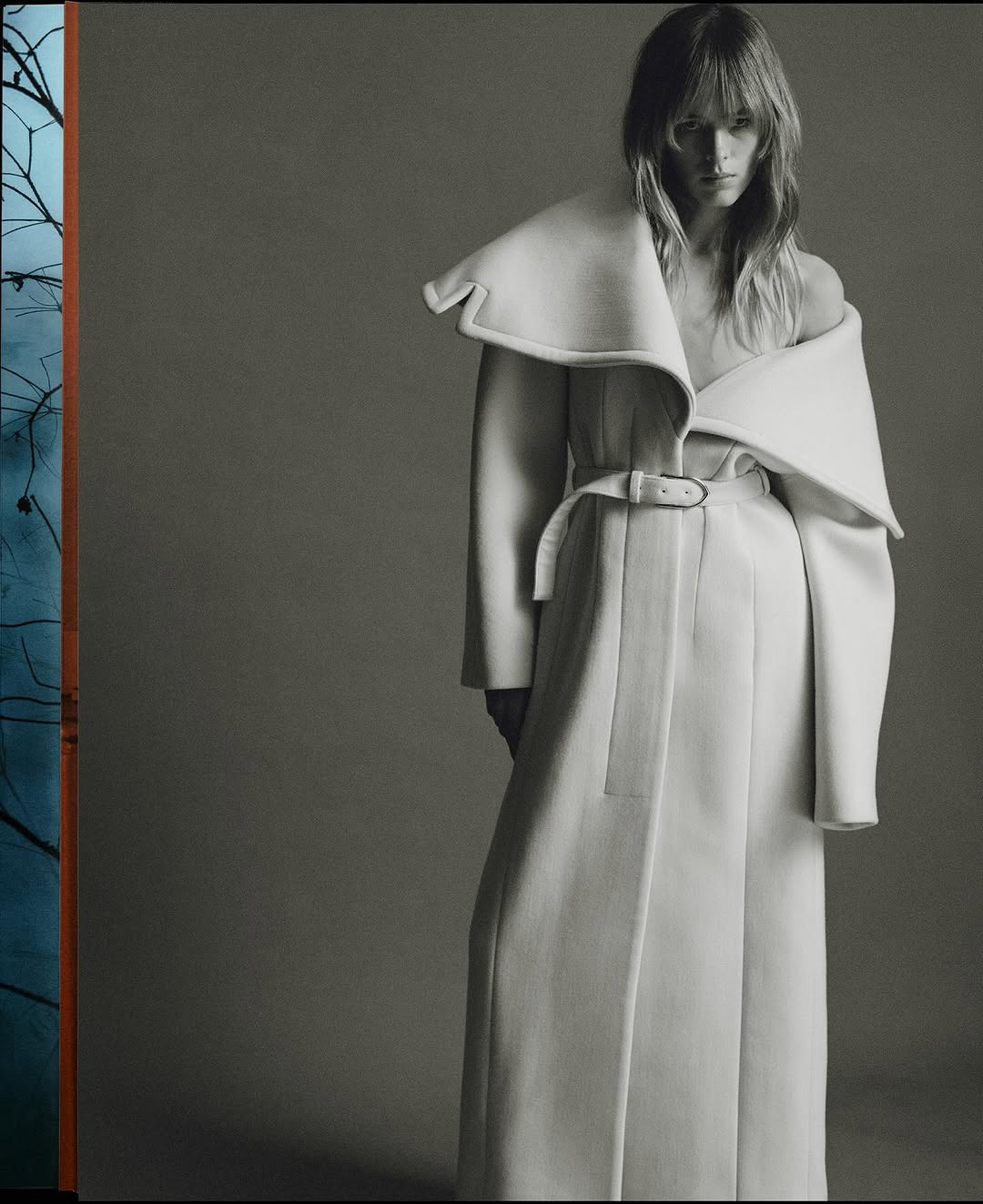 A woman with shoulder-length blonde hair poses in a fashionable floor-length white coat with a belt, shot in a studio setting.