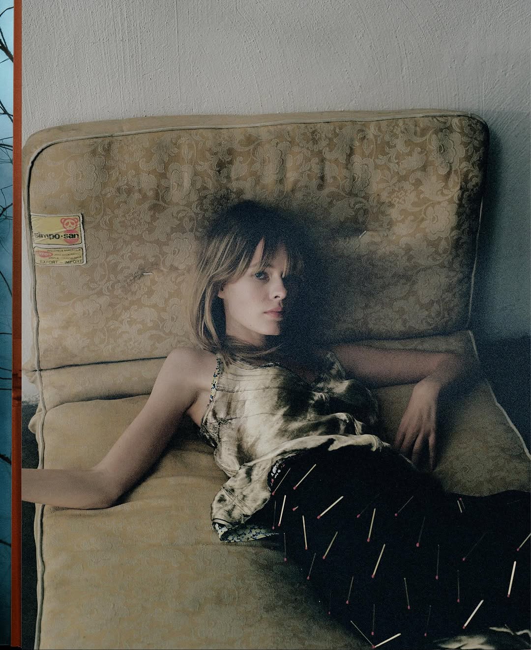 Young woman with blonde hair reclines on patterned, aged upholstered furniture in a close-up studio shot, fashion editorial style.