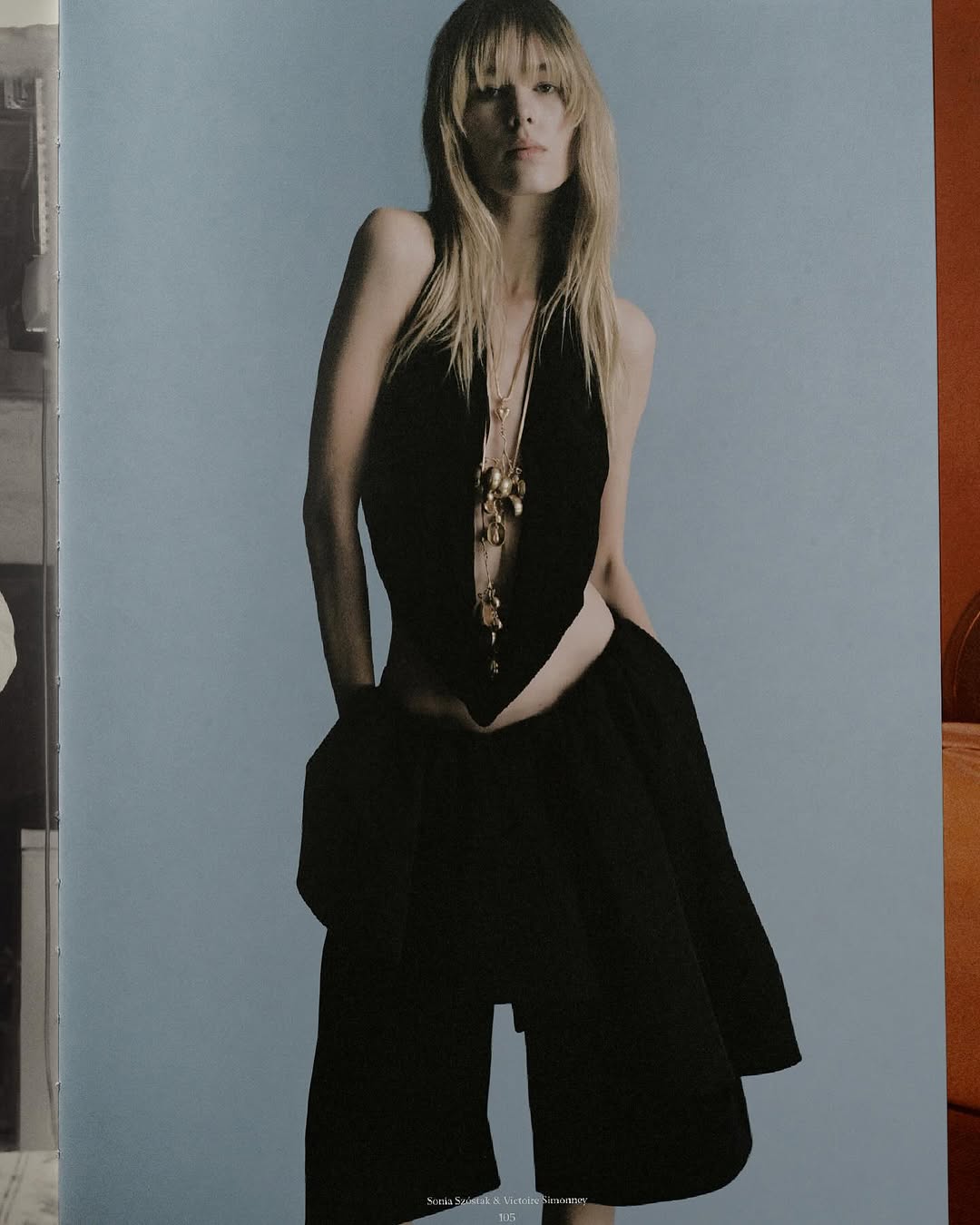 Fashion editorial shot of a model wearing a low-cut black top and black shorts, accessorized with gold jewelry, against a light blue backdrop.