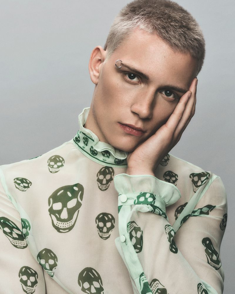 Close-up portrait of a young adult with short light hair wearing a patterned sheer top, posing against a grey background in a studio.
