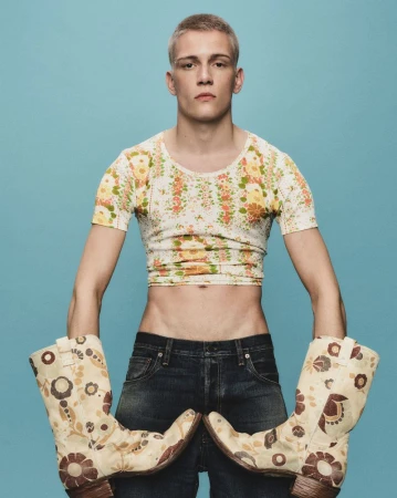 Young male posing with cowboy boots in studio. Man wears cropped floral t-shirt, denim jeans against a light blue seamless backdrop.
