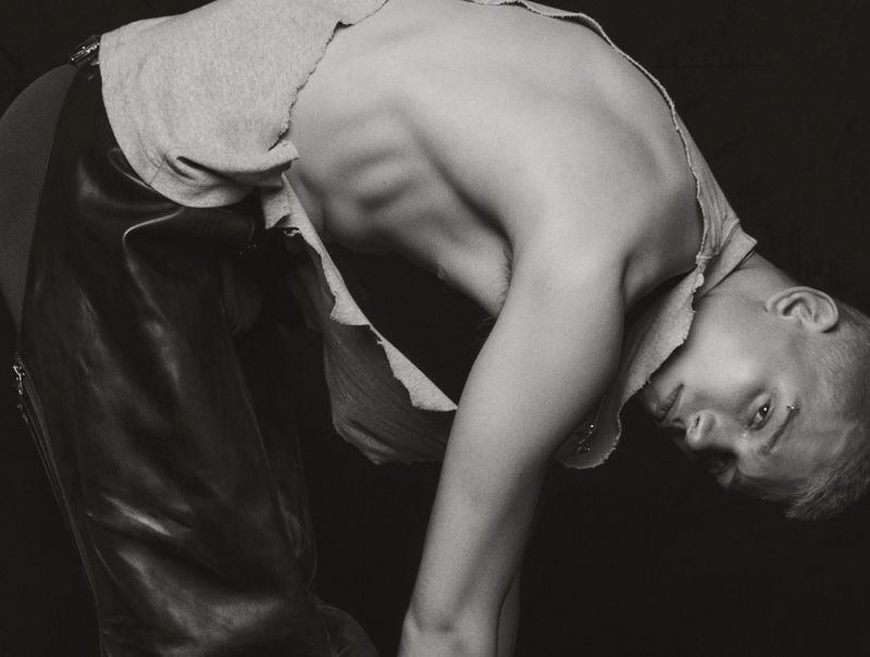 Monochrome portrait of person with short hair, wearing torn gray shirt and leather pants bending over with face in the frame.