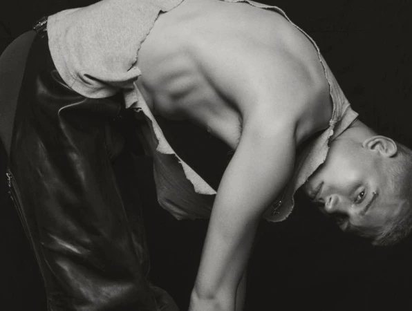 Monochrome portrait of person with short hair, wearing torn gray shirt and leather pants bending over with face in the frame.